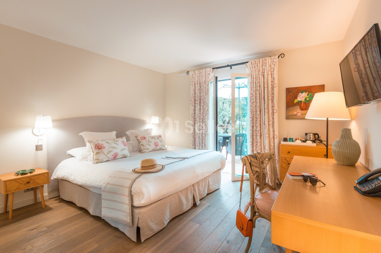 Chambre lumineuse avec lit double, bureau, télévision, et accès à un balcon donnant sur un espace extérieur verdoyant.