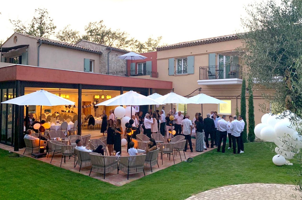 Groupe de personnes rassemblées dans un jardin autour d'une terrasse éclairée, avec des parasols et des ballons blancs.