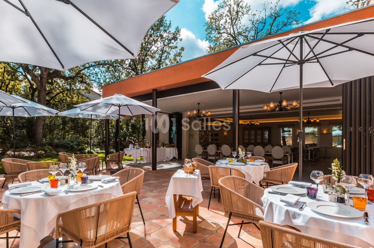 Terrasse d'un restaurant avec tables dressées, chaises en rotin et parasols blancs, entourée de végétation.