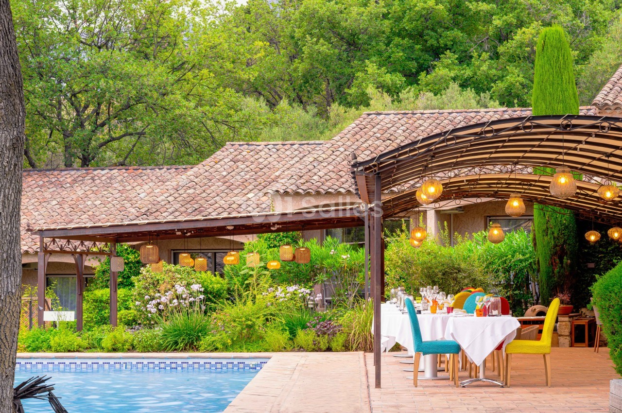 Terrasse avec table dressée près d'une piscine, entourée de végétation et éclairée par des lampes suspendues.