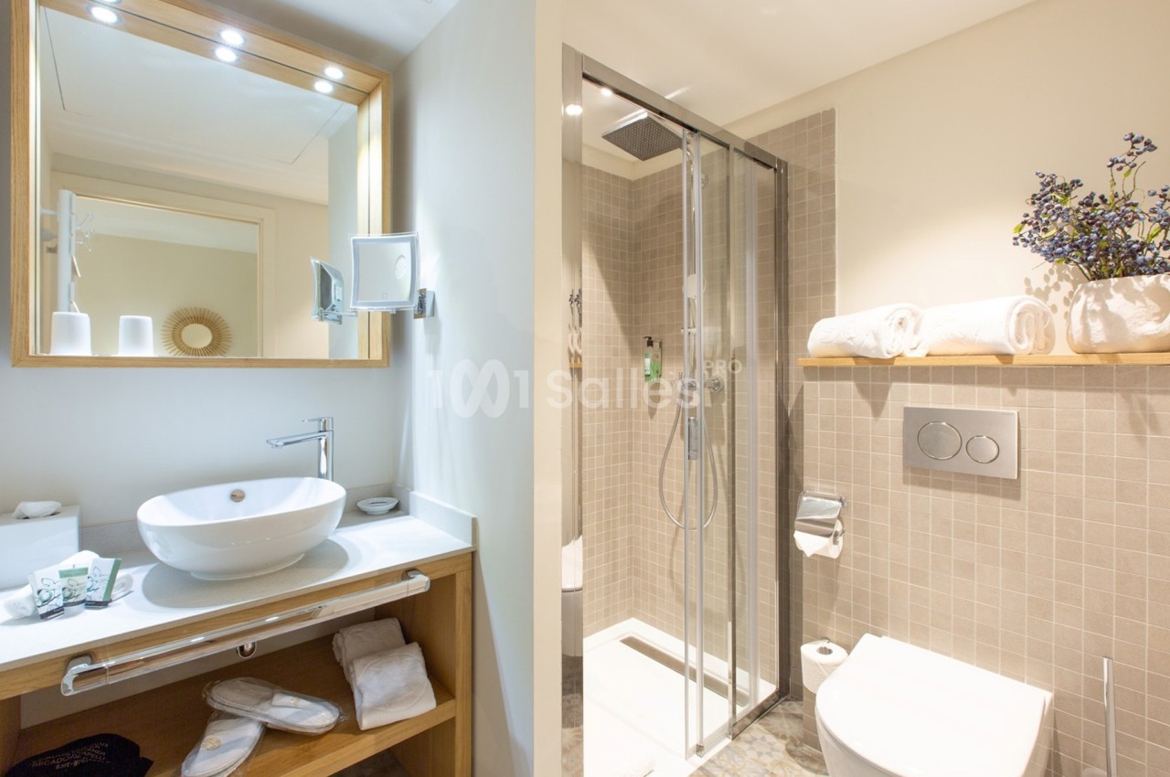 Salle de bain moderne avec douche vitrée, lavabo sur meuble en bois, miroir éclairé et étagère avec serviettes.