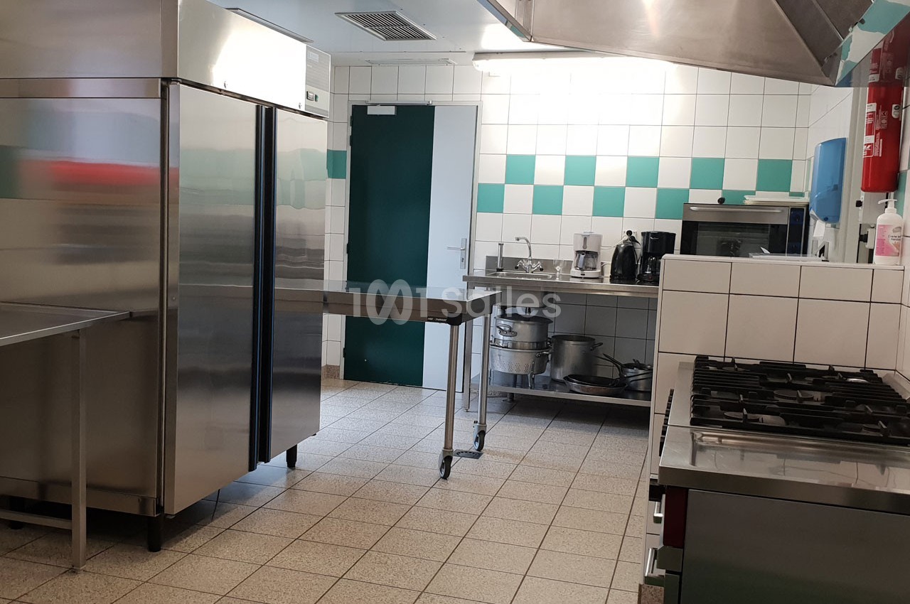 Cuisine professionnelle équipée avec réfrigérateur, plan de travail en inox, évier, plaques de cuisson et carrelage mural.