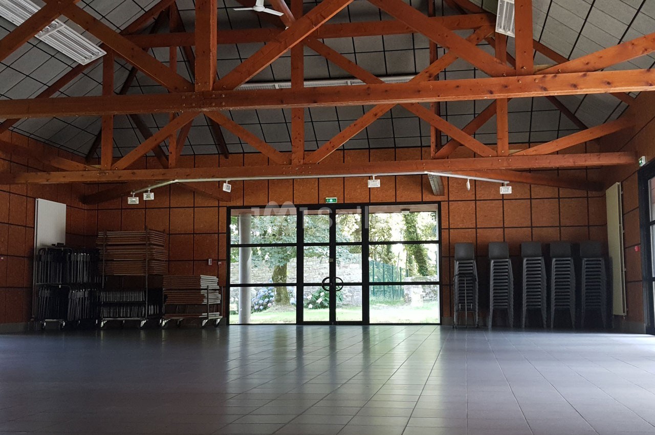 Salle vide avec charpente en bois, sol carrelé, chaises empilées et tables pliées près d'une baie vitrée donnant sur un…