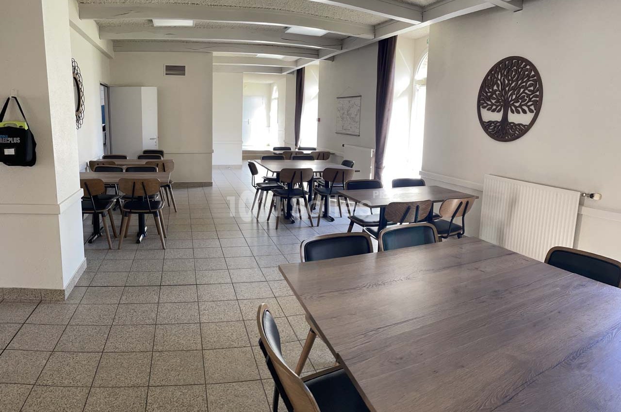 Salle lumineuse avec plusieurs tables et chaises, carrelage au sol, décorations murales et grandes fenêtres avec rideaux.