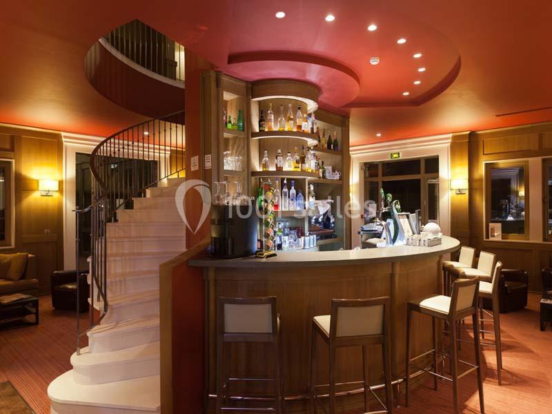 Bar circulaire en bois avec étagères garnies de bouteilles, éclairage chaleureux et escalier en colimaçon à gauche.