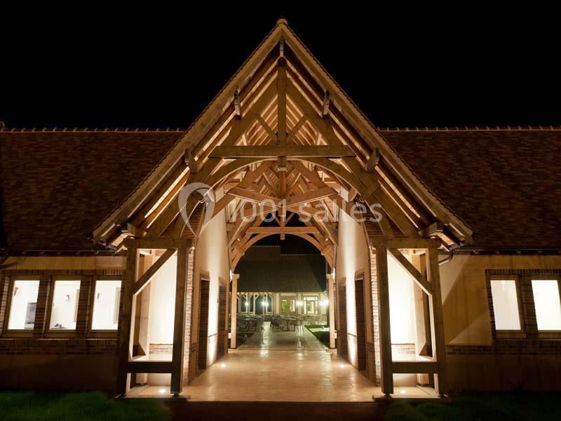 Entrée éclairée d'un bâtiment en bois et briques, avec une structure en charpente apparente, vue de nuit.
