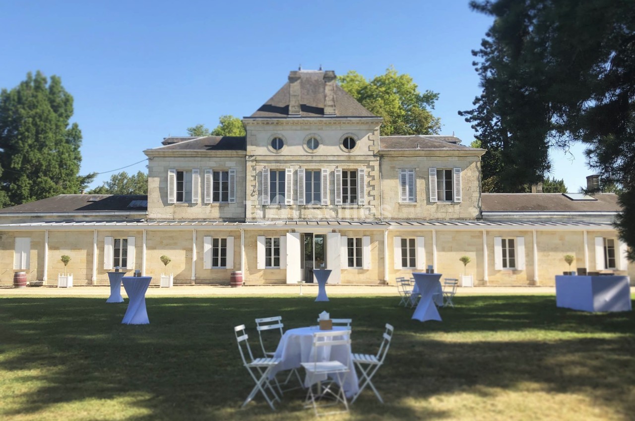 Façade d'un bâtiment en pierre entouré d'arbres, avec des tables et chaises blanches disposées sur une pelouse.