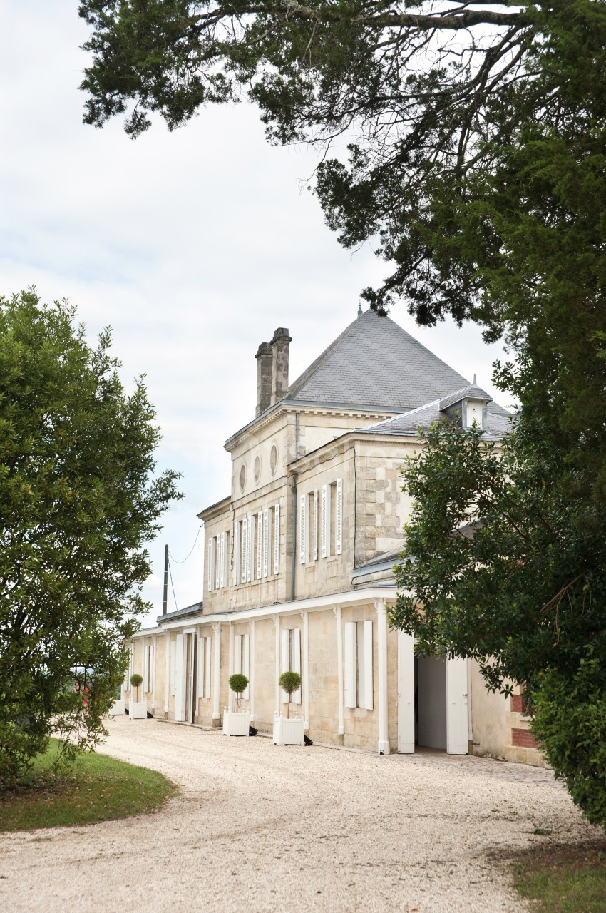Façade d'un bâtiment en pierre à deux étages, entouré d'arbres et bordé d'une allée gravillonnée.