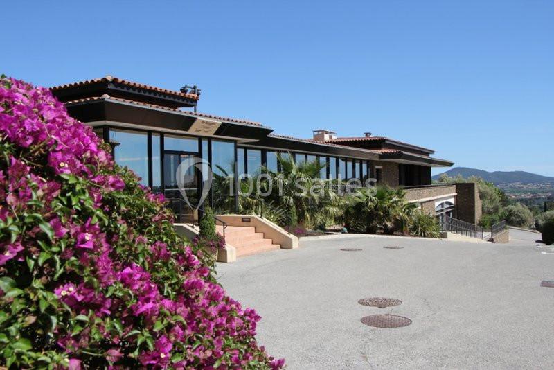 Bâtiment moderne avec grandes baies vitrées, entouré de bougainvilliers et de palmiers, sous un ciel bleu clair.