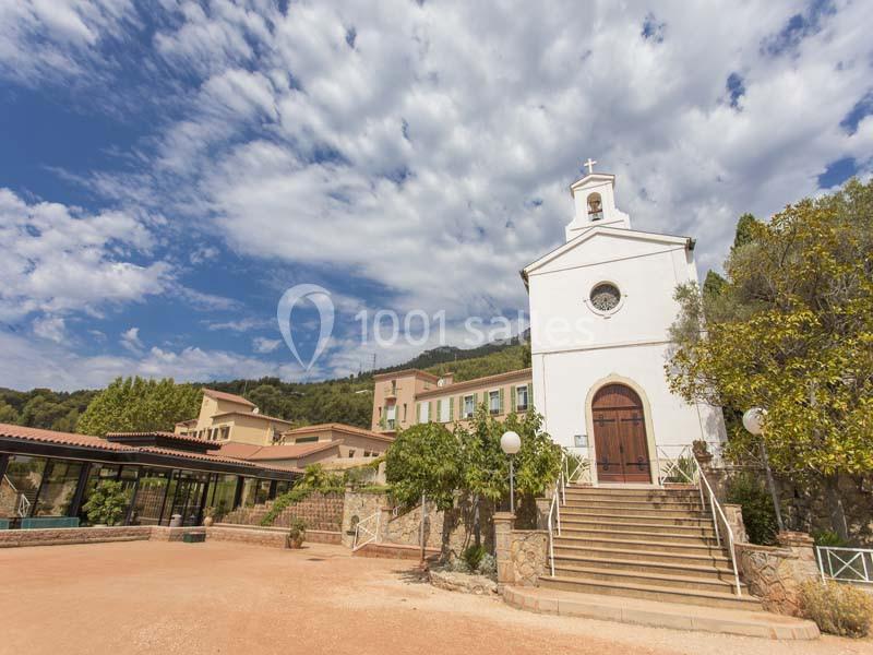 Église blanche avec clocher, entourée de bâtiments et de végétation, sous un ciel partiellement nuageux.
