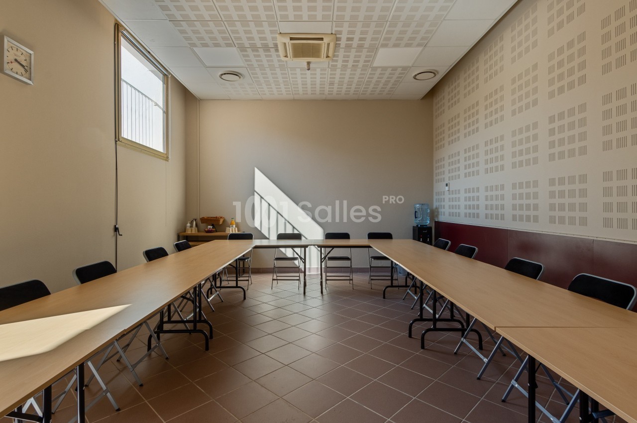 Salle de réunion lumineuse avec tables disposées en U, chaises noires, fenêtre, horloge murale et fontaine à eau.