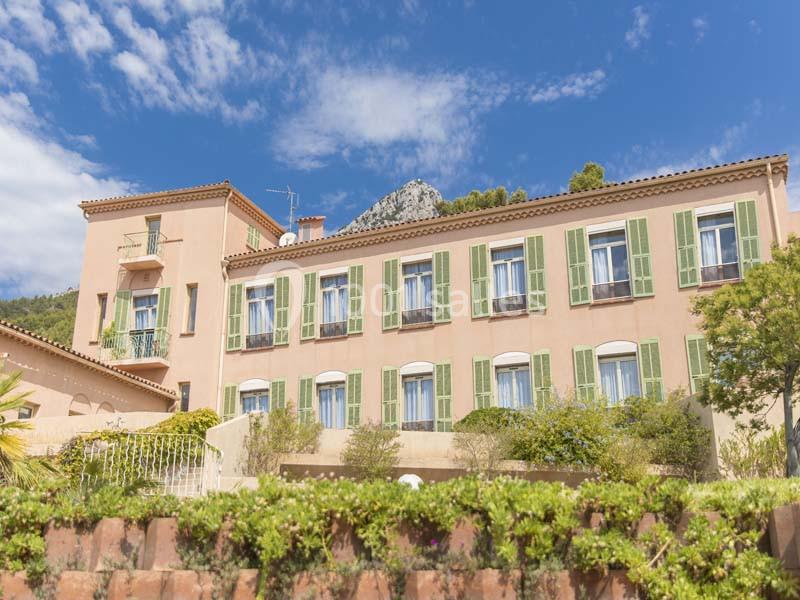 Bâtiment méditerranéen rose avec volets verts, entouré de végétation, sous un ciel partiellement nuageux.