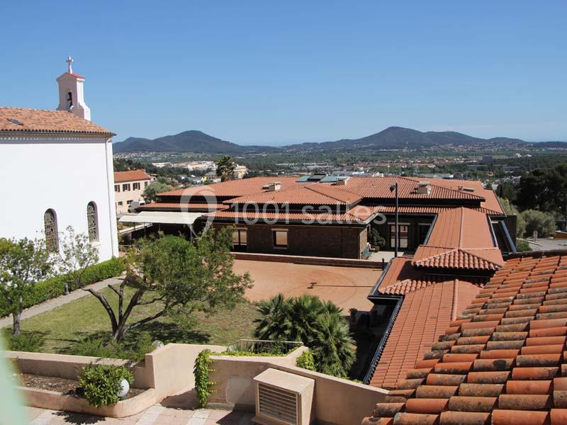 Vue d'un bâtiment à toits en tuiles entouré de végétation, avec une église et des collines à l'arrière-plan.