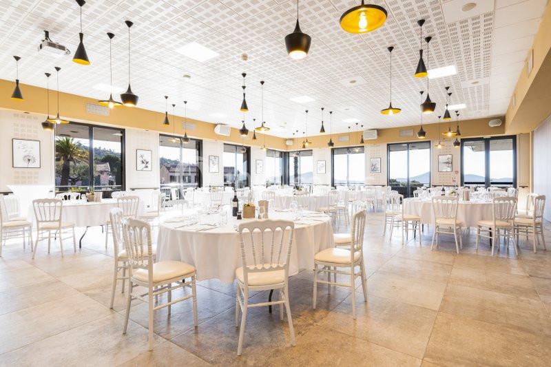 Salle lumineuse avec tables rondes dressées, chaises blanches et grandes baies vitrées donnant sur un paysage extérieur.