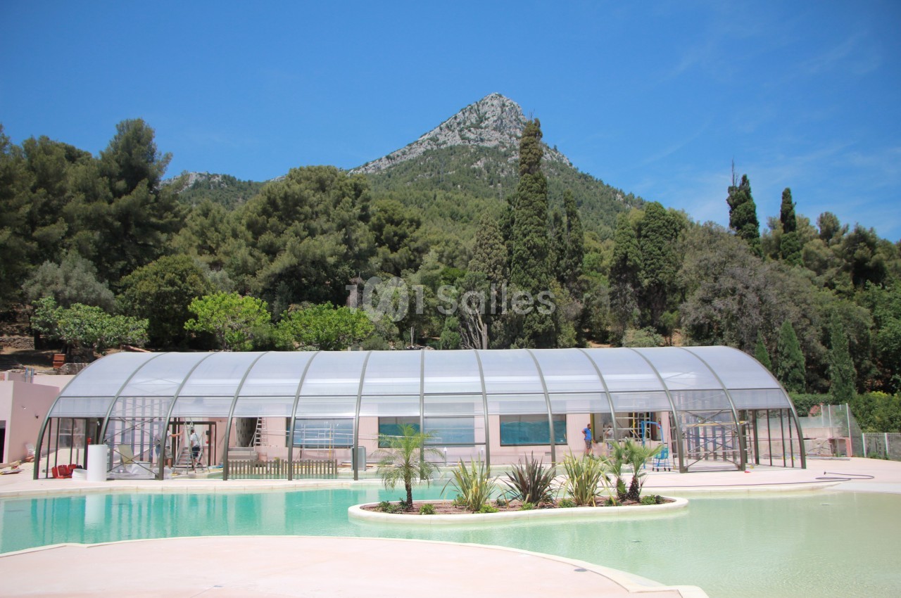 Piscine extérieure avec abri transparent, entourée de végétation et située au pied d'une colline boisée.
