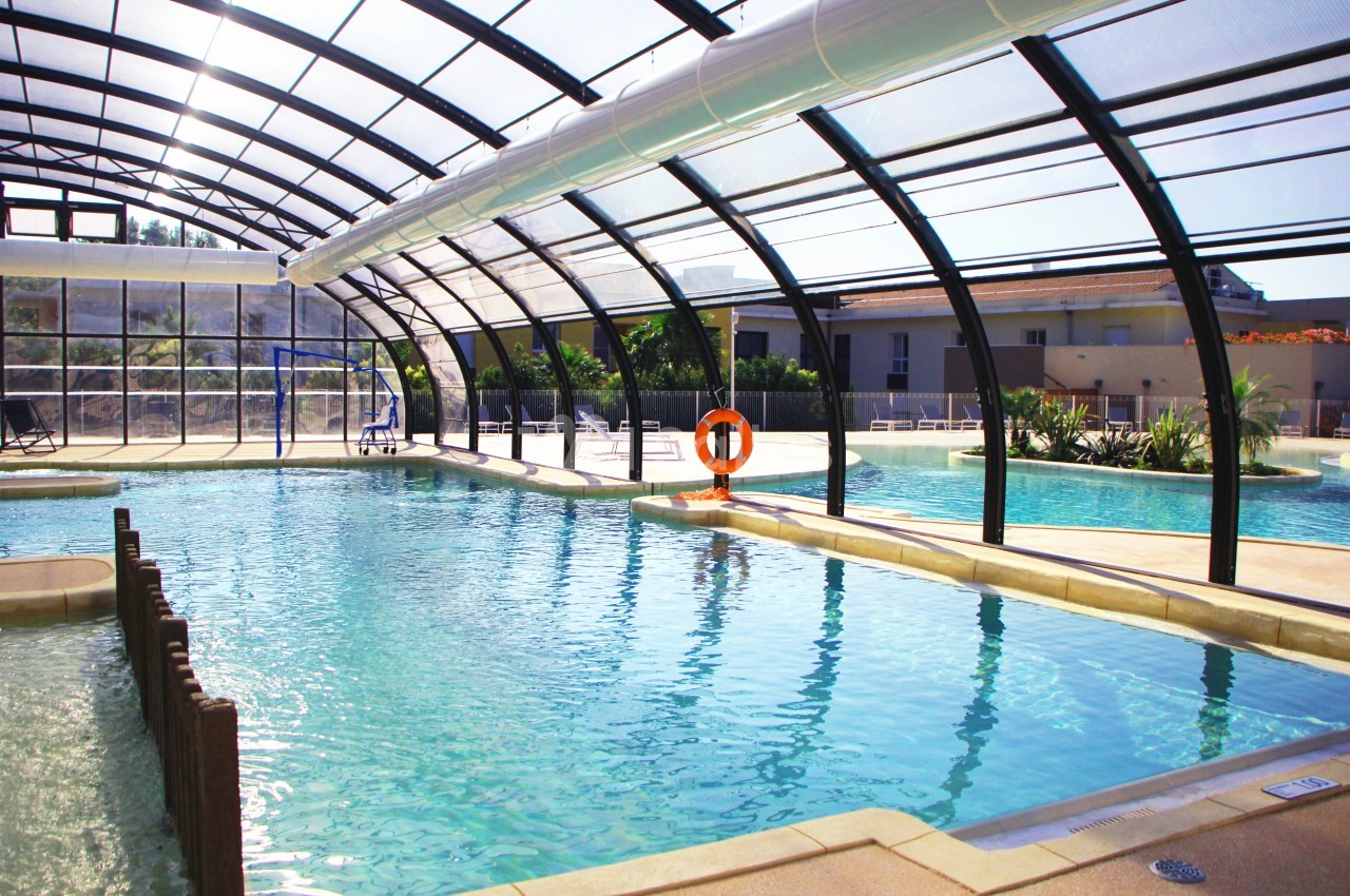 Piscine intérieure avec toit transparent, séparations en bois et bouée de sauvetage orange accrochée au fond.