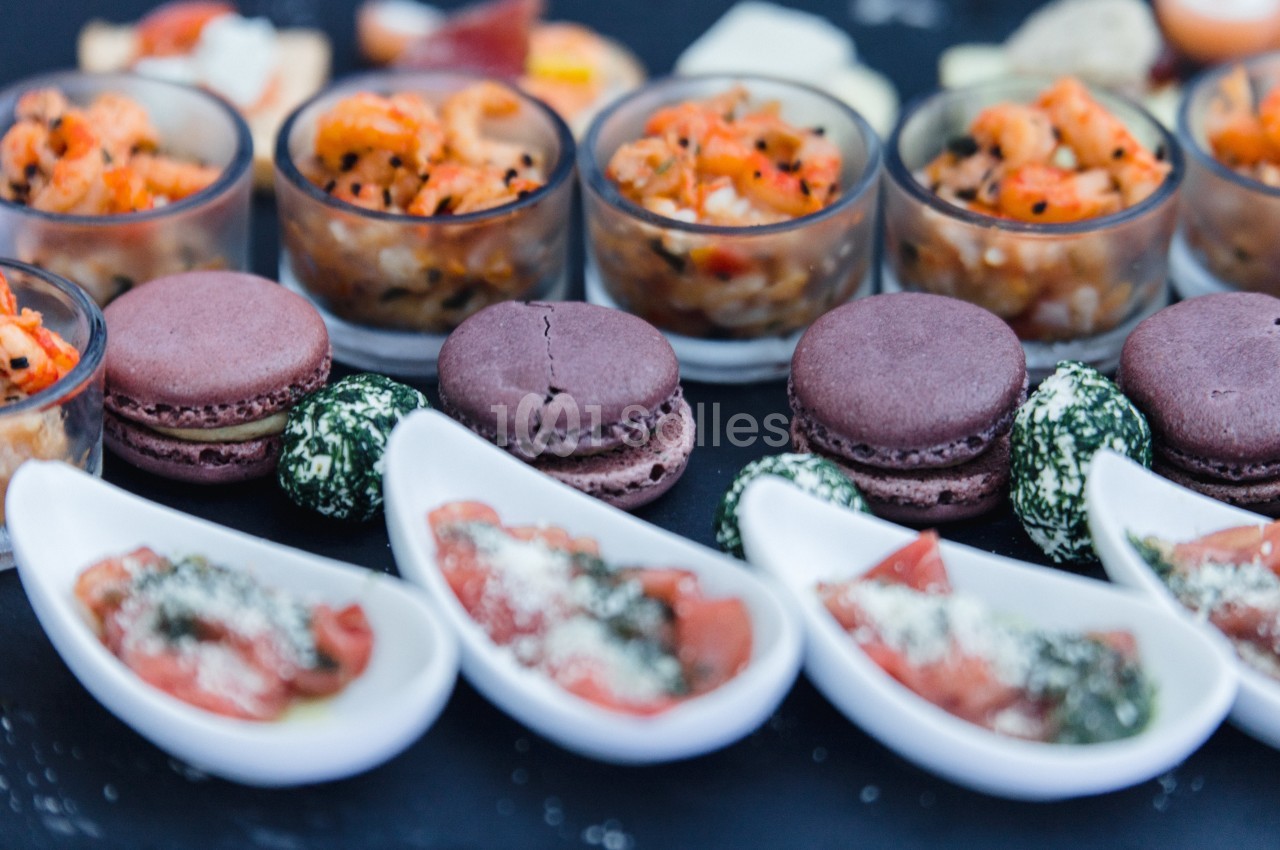 Assortiment de verrines, macarons salés, bouchées de fromage et cuillères apéritives sur une table noire.