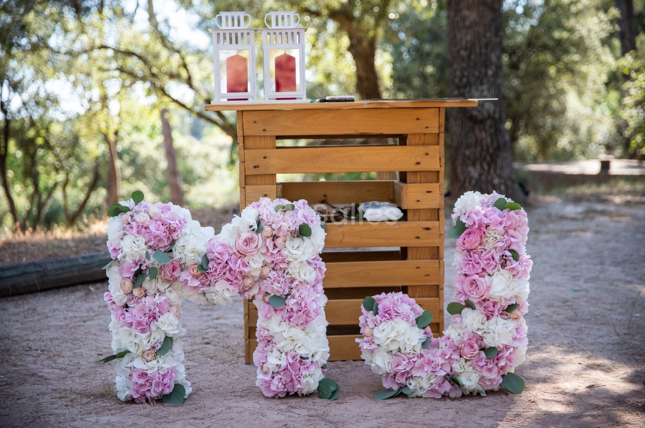 Décoration extérieure avec des lettres ’M’ et ’W’ ornées de fleurs roses et blanches devant une table en bois.