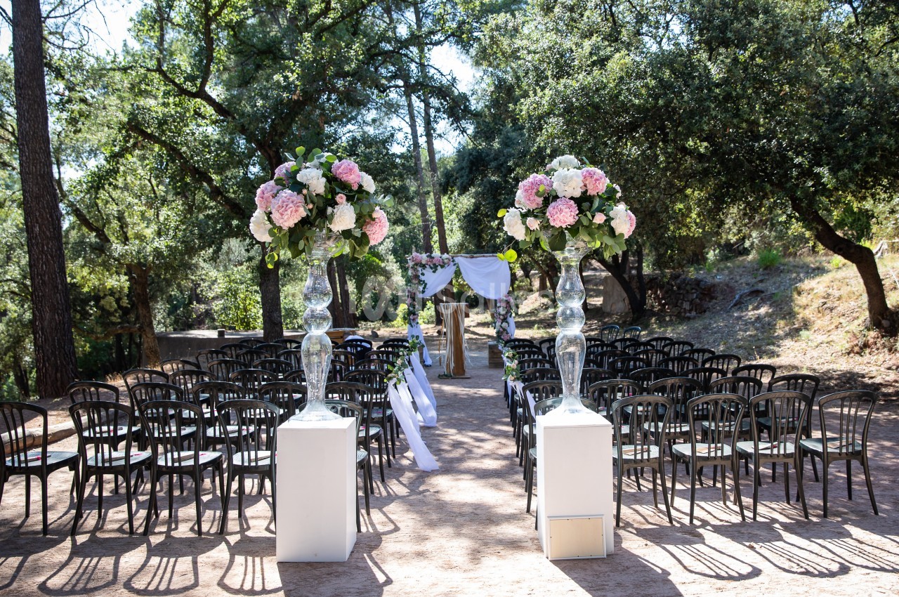 Allée centrale décorée de fleurs menant à une arche nuptiale dans un cadre extérieur boisé.