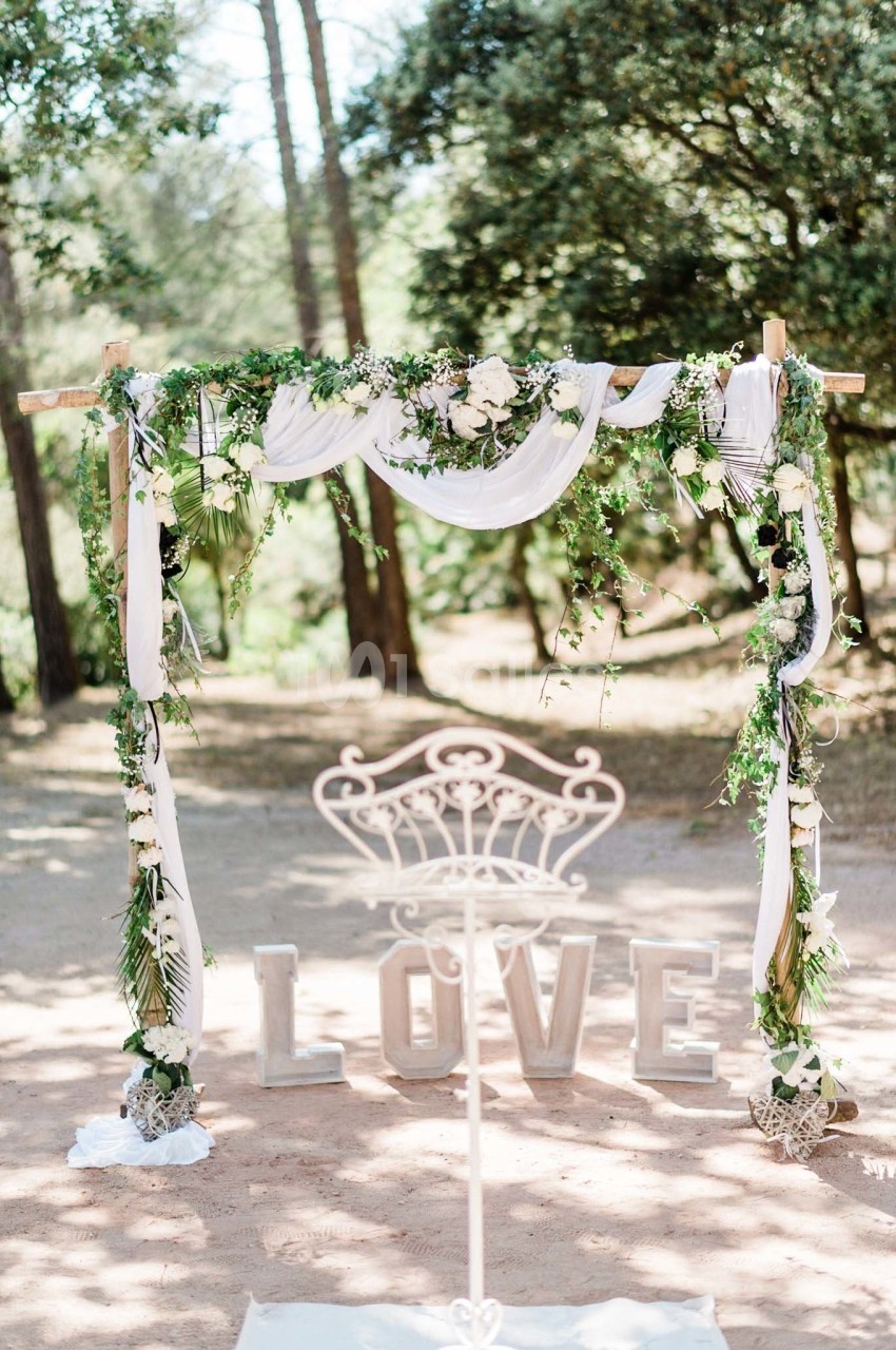 Arche de mariage décorée de fleurs, feuillages et tissus blancs, avec des lettres formant le mot ’LOVE’ au sol.