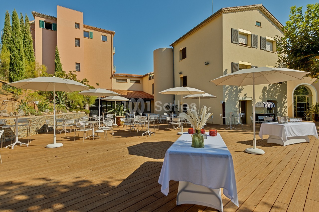 Terrasse en bois avec tables, chaises et parasols blancs, entourée de bâtiments colorés et de végétation.