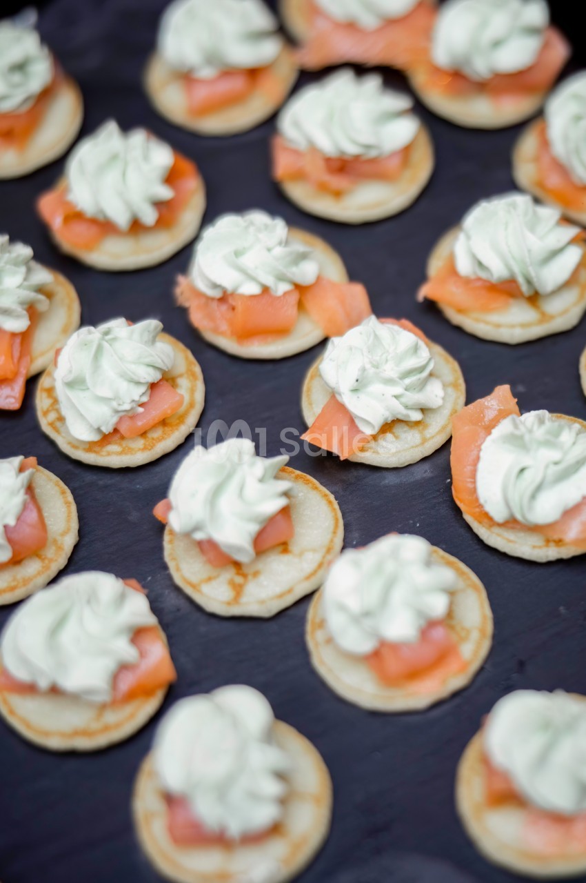 Petits blinis garnis de crème fouettée et de tranches de saumon fumé, disposés sur une surface sombre.