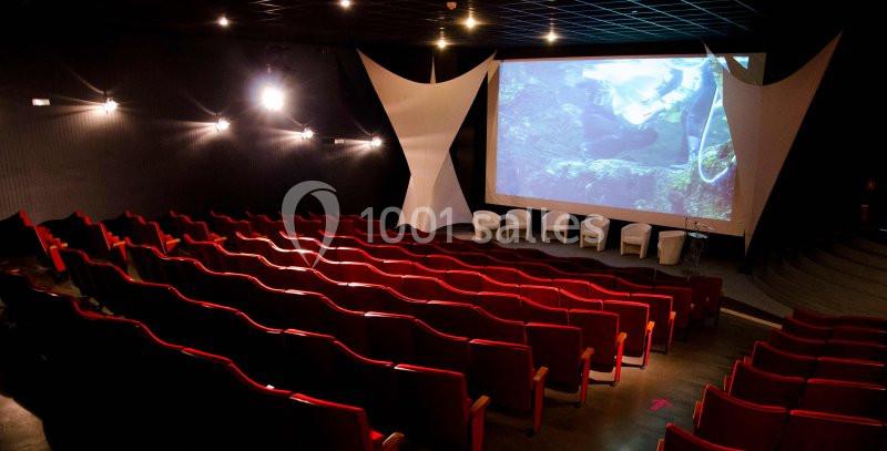 Salle de projection avec des rangées de fauteuils rouges, un écran allumé et des chaises blanches sur la scène.