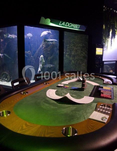 Table de jeu de casino avec cartes et jetons, située devant un grand aquarium éclairé.