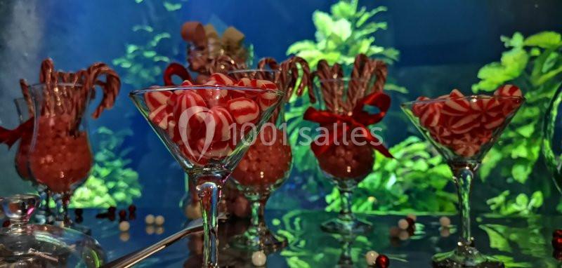 Verres et vases remplis de bonbons rouges et blancs, décorés de rubans rouges, devant un fond végétal lumineux.