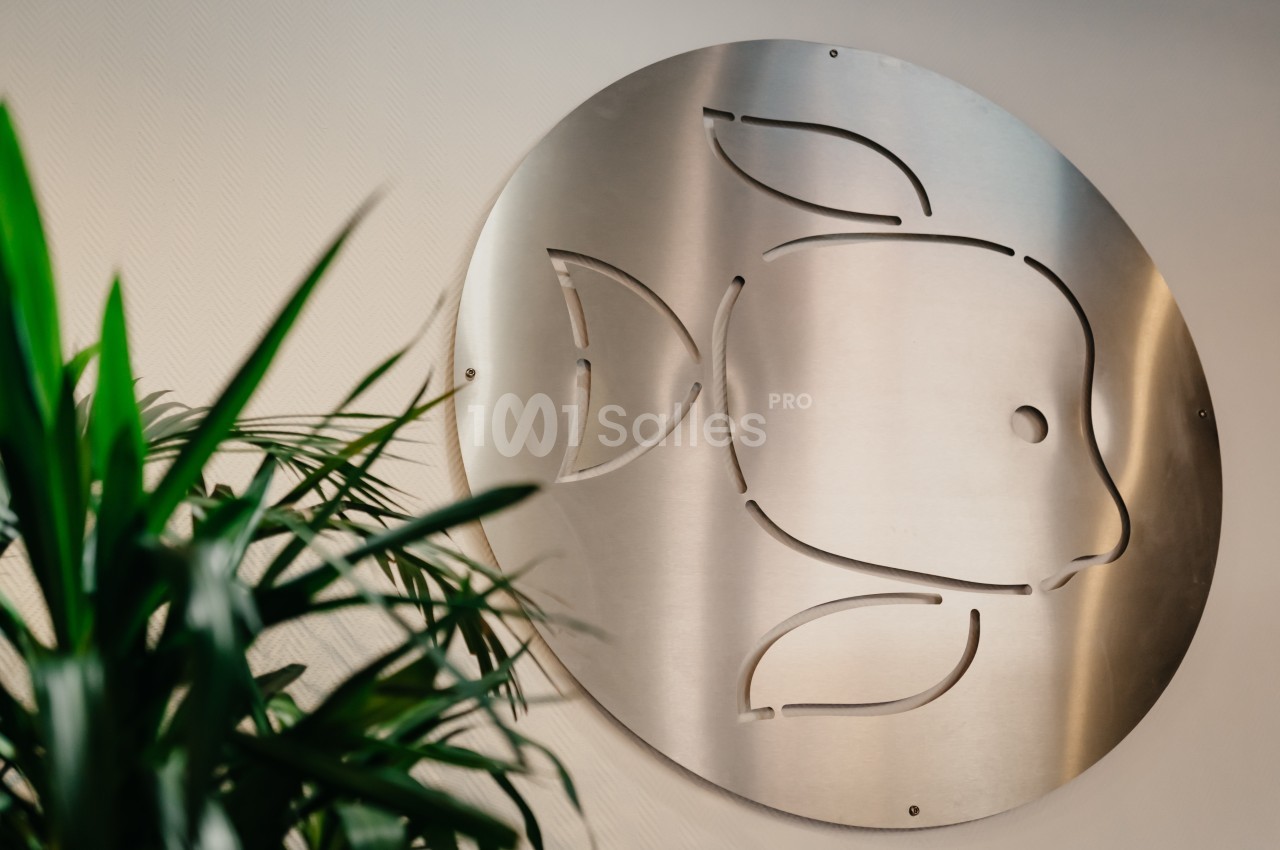 Plaque métallique ronde avec un motif de poisson découpé, accrochée à un mur clair, partiellement cachée par une plante…