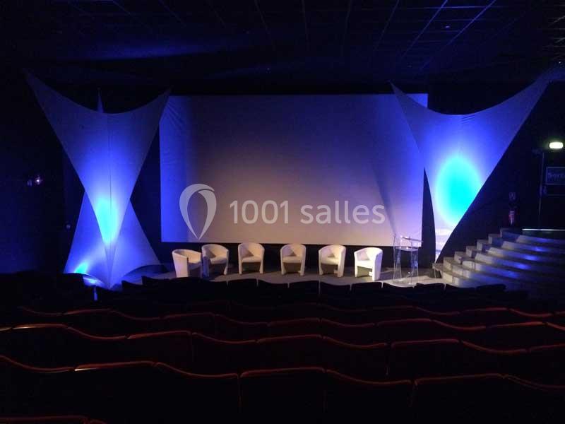 Salle de conférence avec écran de projection, chaises blanches alignées et éclairage bleu sur des structures décoratives.