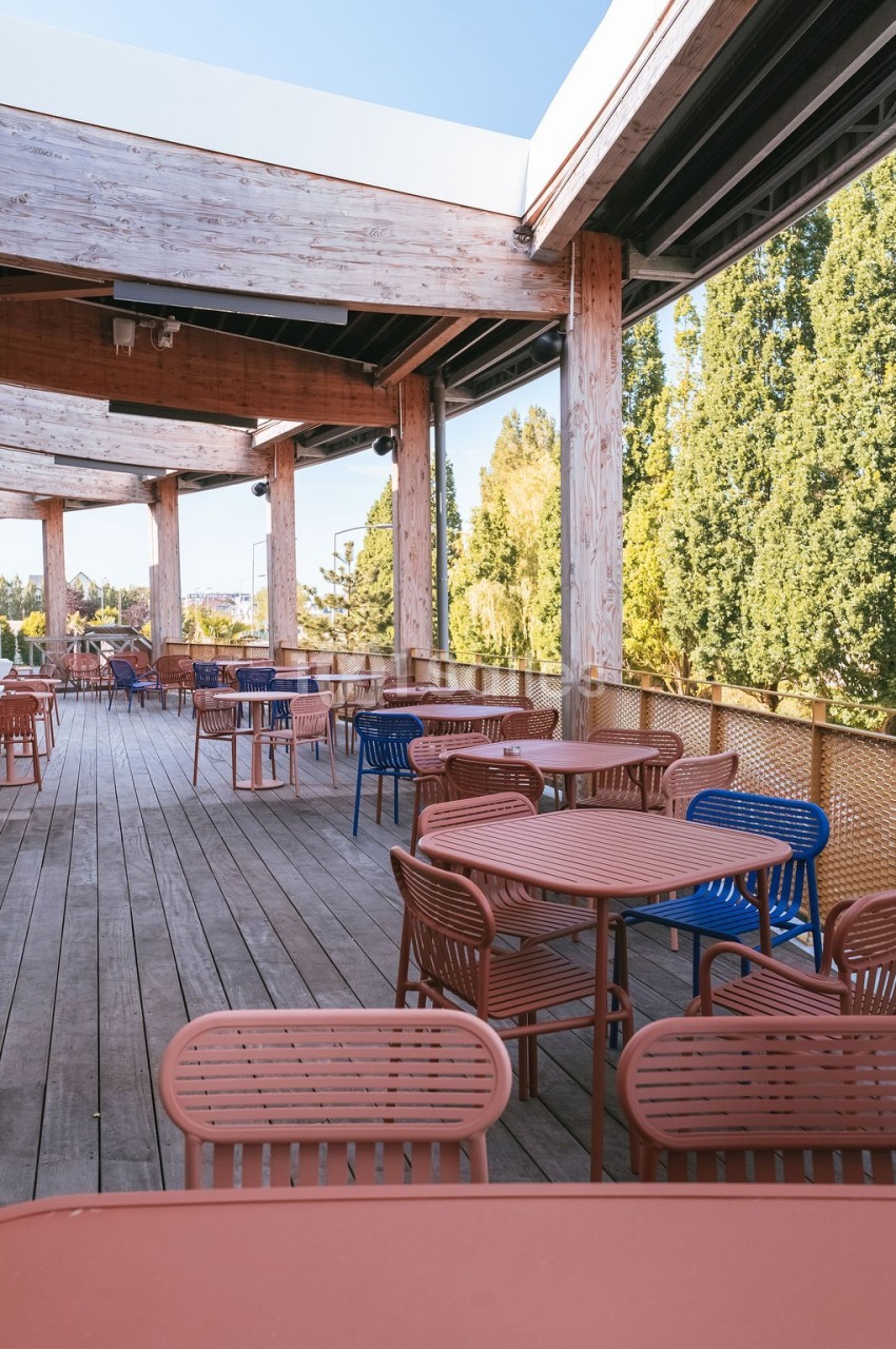 Terrasse en bois avec des tables et chaises colorées, entourée d'arbres et partiellement couverte par une structure en bois.