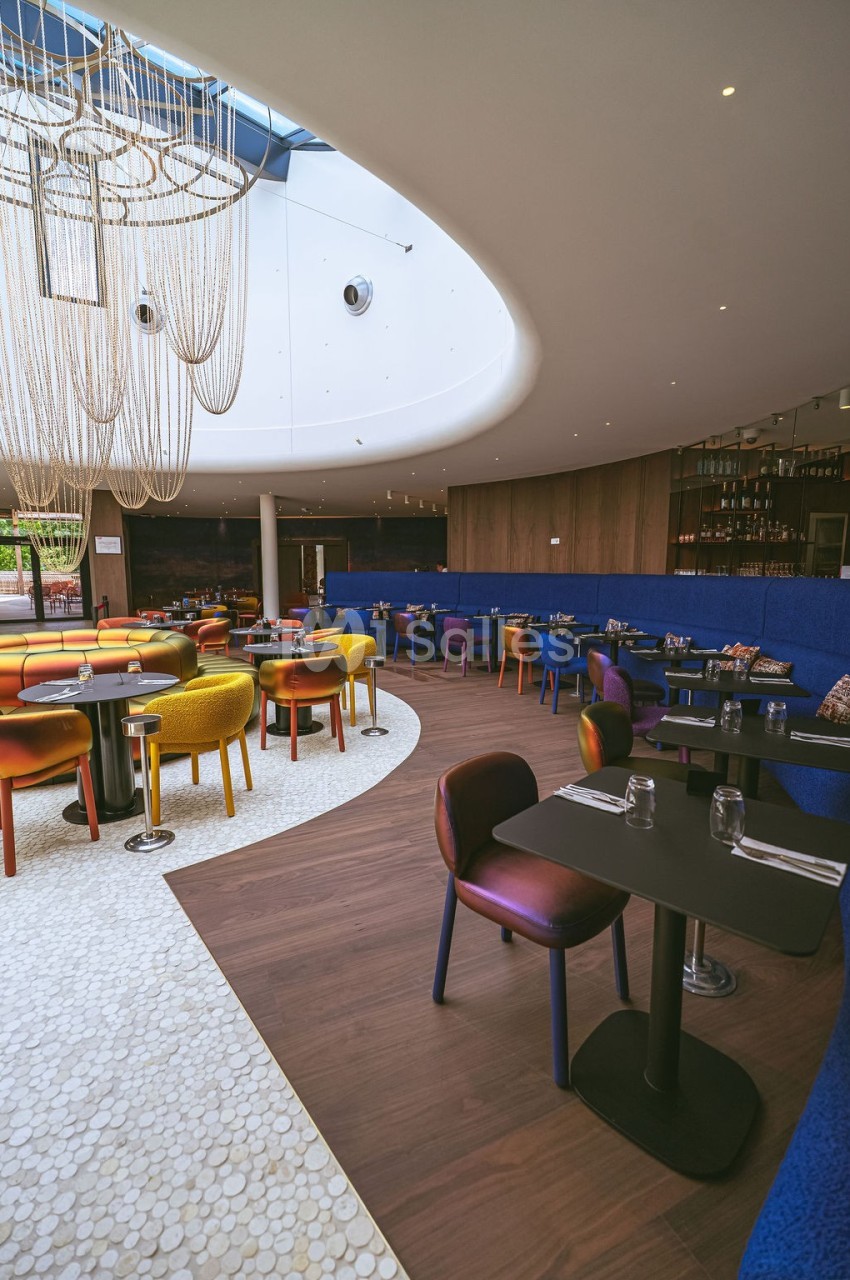 Salle de restaurant moderne avec chaises colorées, tables dressées et grand lustre suspendu sous une verrière.