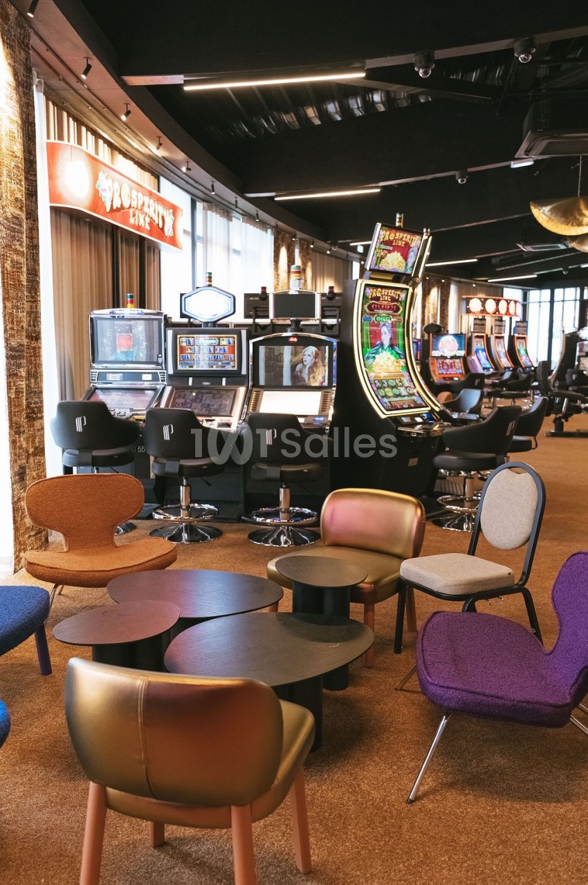 Salle de jeux avec machines à sous alignées et espace détente aménagé avec des fauteuils colorés et des tables basses.