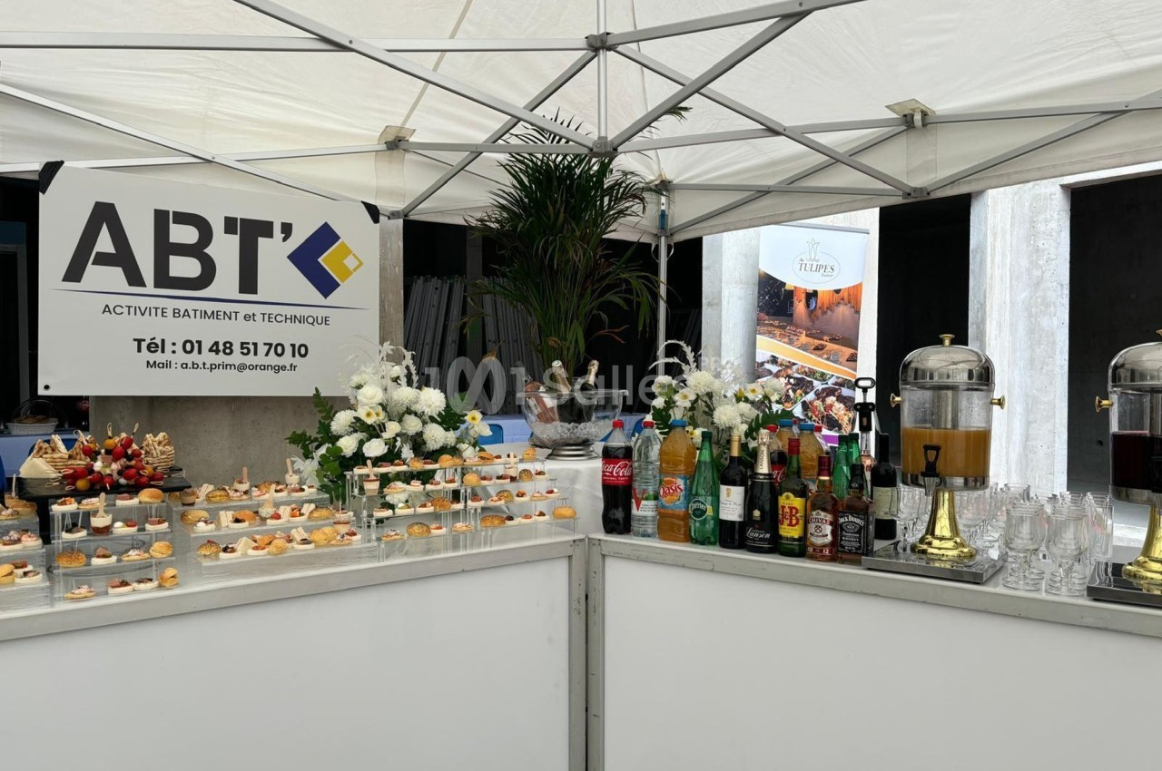 Stand sous une tente avec buffet de boissons et amuse-bouches, décoré de fleurs blanches et affiches promotionnelles.