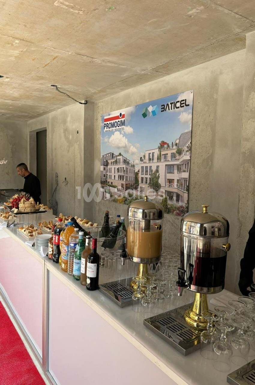 Buffet avec boissons, viennoiseries et fruits, installé dans une salle en béton brut avec affiches de projets immobiliers.