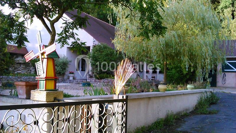 Vue d'une maison avec jardin, clôture en métal, arbre pleureur et moulin décoratif coloré au premier plan.