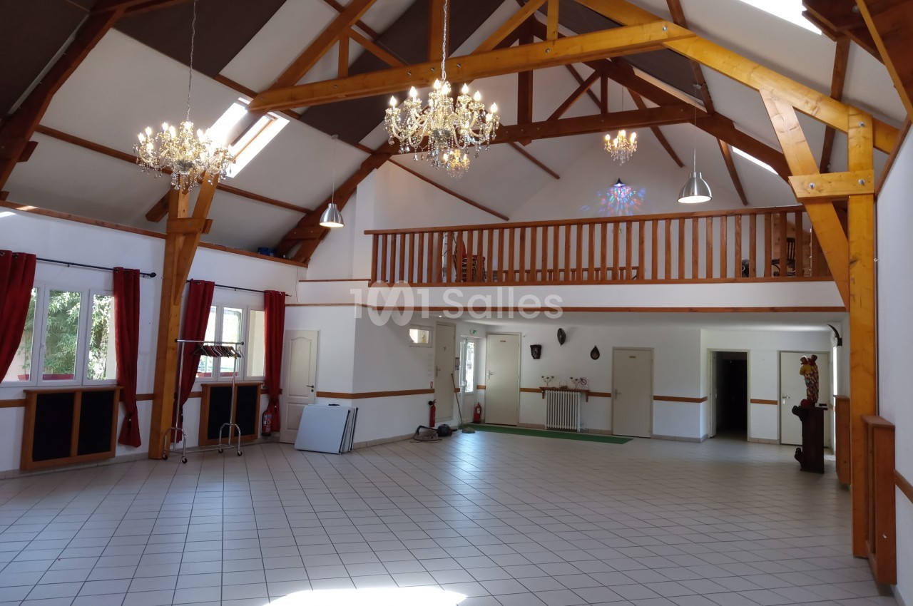 Grande salle lumineuse avec poutres apparentes, lustres, mezzanine en bois et carrelage au sol.
