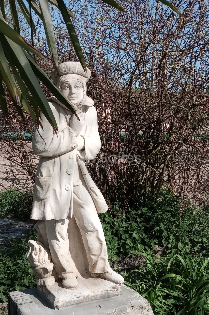Statue en pierre représentant une personne en manteau et chapeau, entourée de végétation et éclairée par le soleil.
