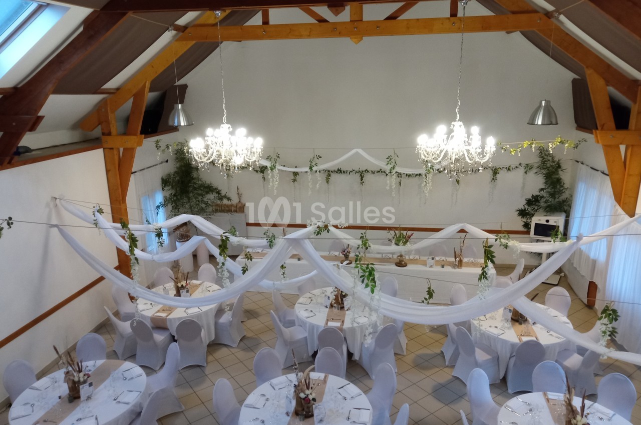 Salle de réception décorée pour un événement, avec tables rondes, nappes blanches, guirlandes et chandeliers suspendus.