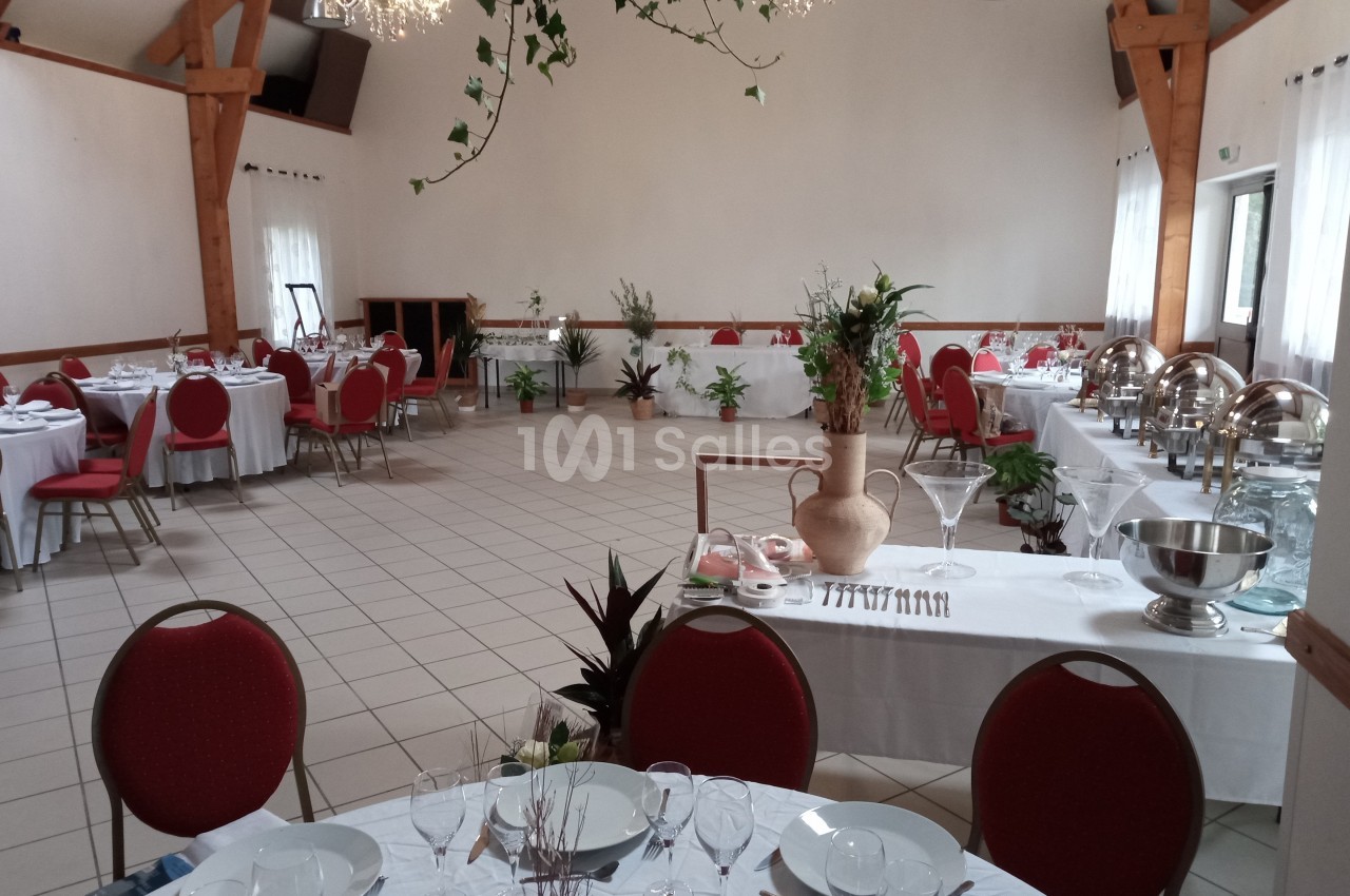 Salle de réception lumineuse avec tables dressées, chaises rouges, buffets en inox et décorations végétales.