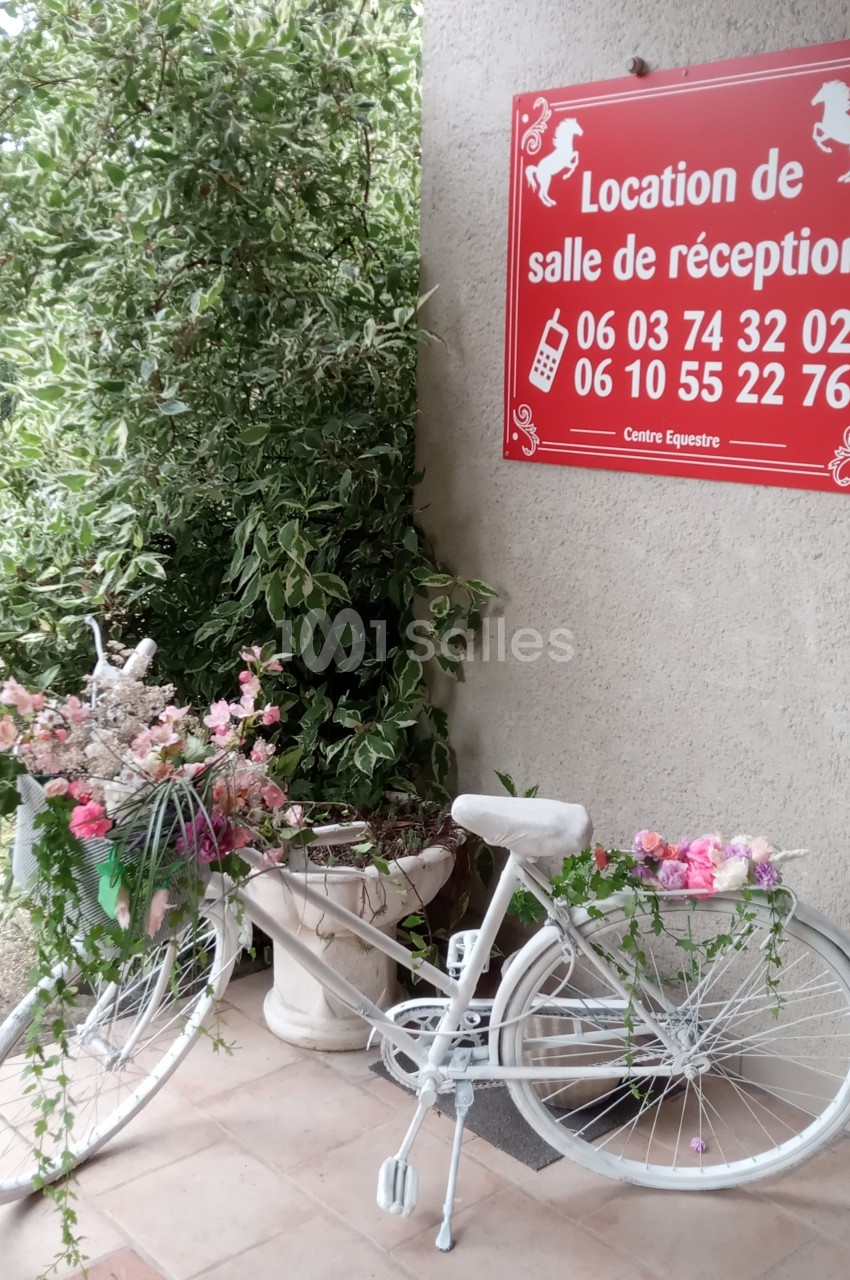 Vélo blanc décoré de fleurs devant un panneau rouge indiquant la location d'une salle de réception.