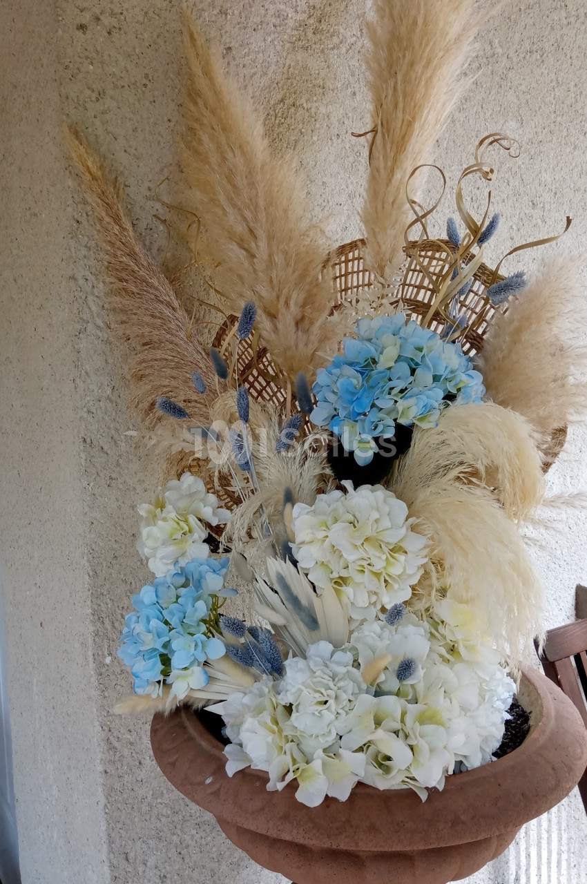 Composition florale avec des fleurs blanches et bleues, pampas et éléments décoratifs dans un vase en terre cuite.
