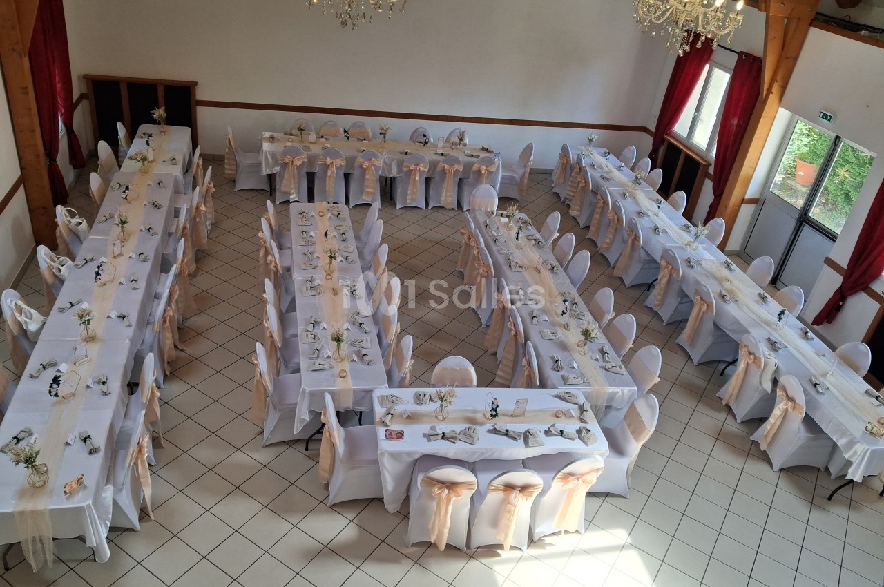 Salle de réception avec tables rectangulaires dressées, nappes blanches et chaises décorées de nœuds beiges.