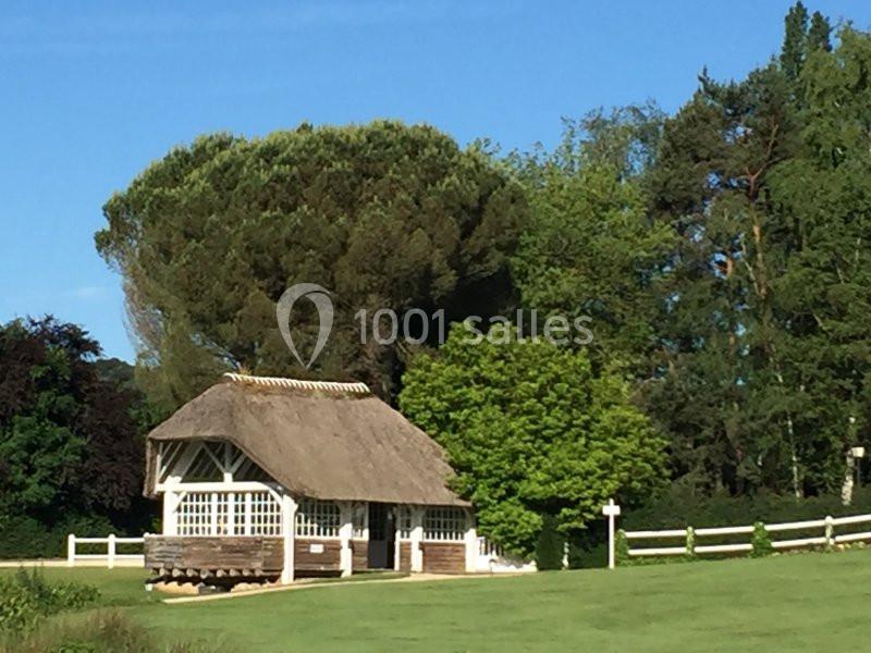 Maison en toit de chaume entourée de verdure, avec des arbres et une clôture blanche en arrière-plan.