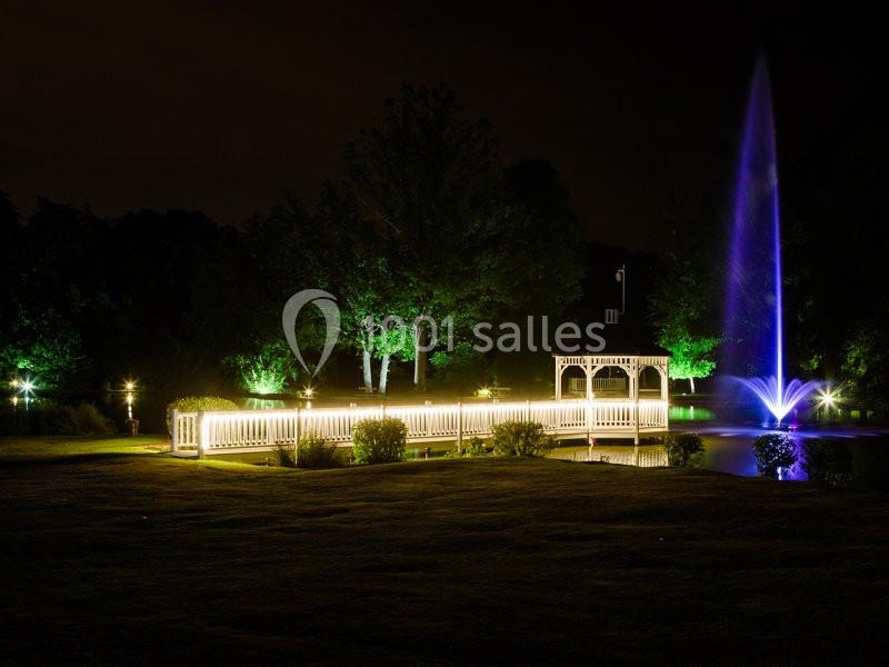 Pont éclairé et kiosque en bois près d'une fontaine illuminée, entourés d'arbres dans un parc nocturne.