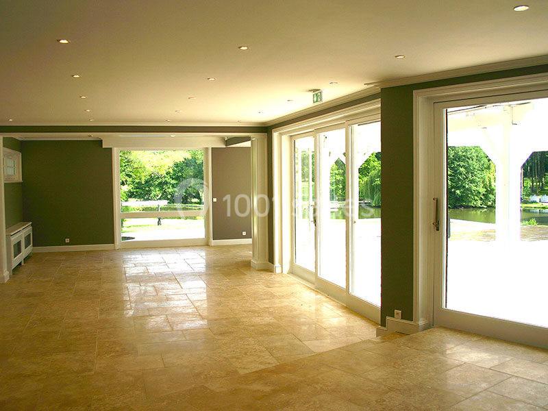 Salle lumineuse avec de grandes baies vitrées donnant sur un jardin verdoyant, sol en carrelage beige.