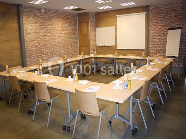 Salle de réunion avec tables disposées en U, chaises en bois, tableau blanc, écran de projection et bouteilles d'eau.