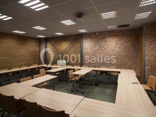 Salle de réunion avec tables en bois disposées en U, chaises assorties et murs en briques et bois.