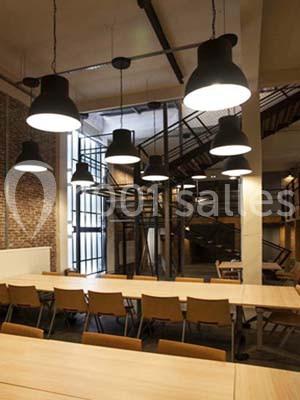 Salle moderne avec de longues tables en bois, chaises alignées, luminaires suspendus et escalier métallique en arrière-plan.